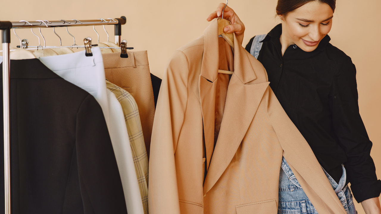 A woman in a black shirt and overalls examines a beige blazer on a clothing rack with various jackets and pants, capturing the essence of effortless French style against a beige background.